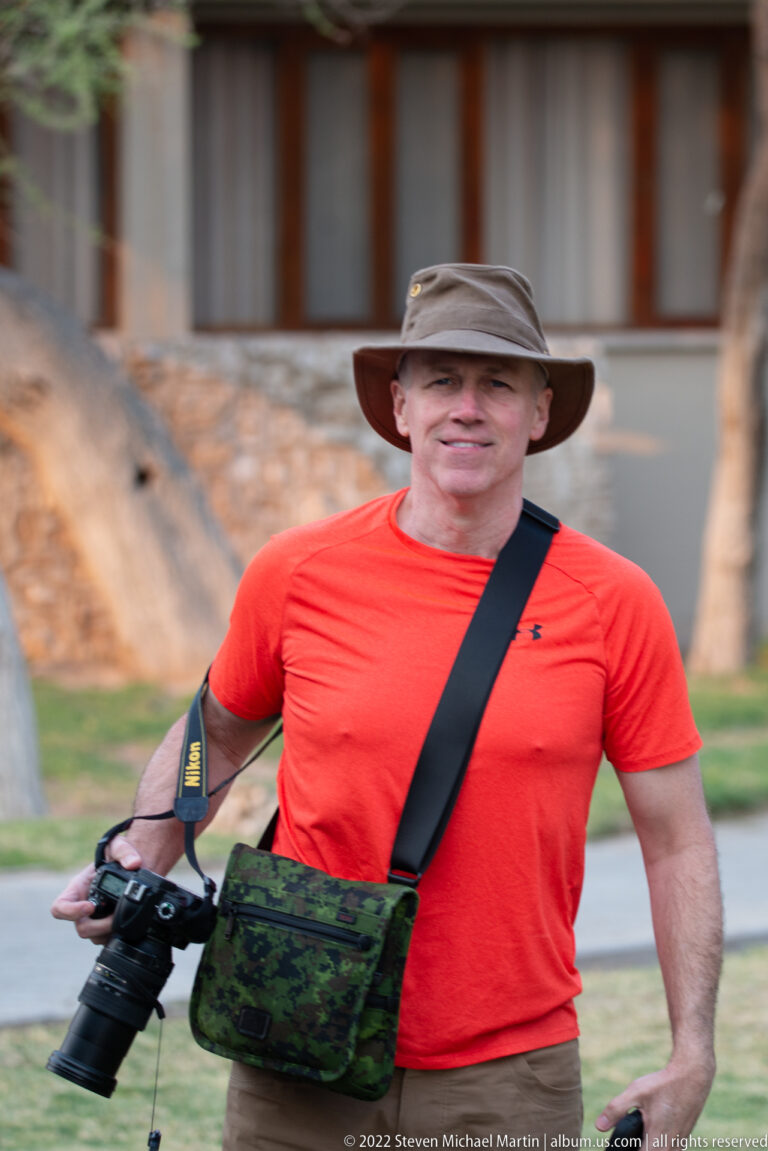 Traveler in red shirt and field hat holding a DSLR with long lens and wearing a camera bag.