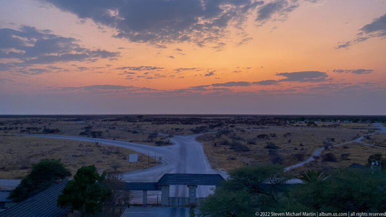 Elevated dawn view over Okaukuejo roofs toward branching roads and flat scrubland beneath a pastel sky.