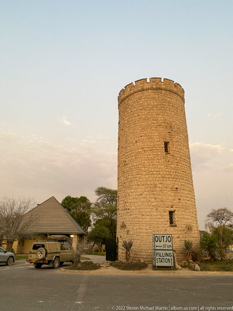 Round stone tower beside an Outjo and filling-station sign, with a safari vehicle parked nearby.