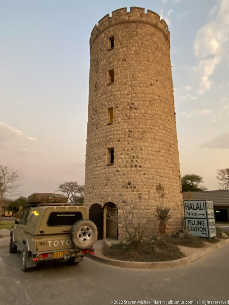 Close view of Okaukuejo's round stone tower with arched doorway, safari vehicle, and Halali direction sign.
