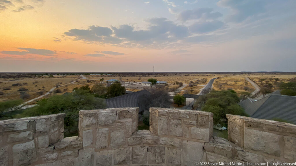 Stone crenellations in foreground overlooking Okaukuejo camp, roads, trees, and flat Etosha plains.