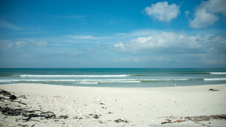 Wide white-sand beach with kelp in the foreground, low surf, turquoise water, and scattered clouds.