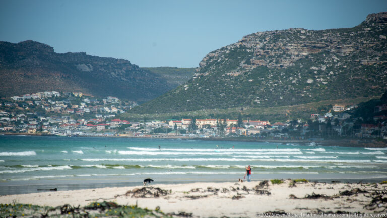 Person walking on a sandy False Bay beach, with breaking waves, hillside homes, and rugged mountains beyond.