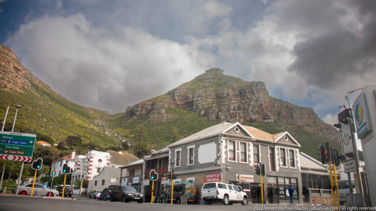 Muizenberg street with historic low-rise buildings, cars, traffic lights, and a steep cloud-covered mountain behind.