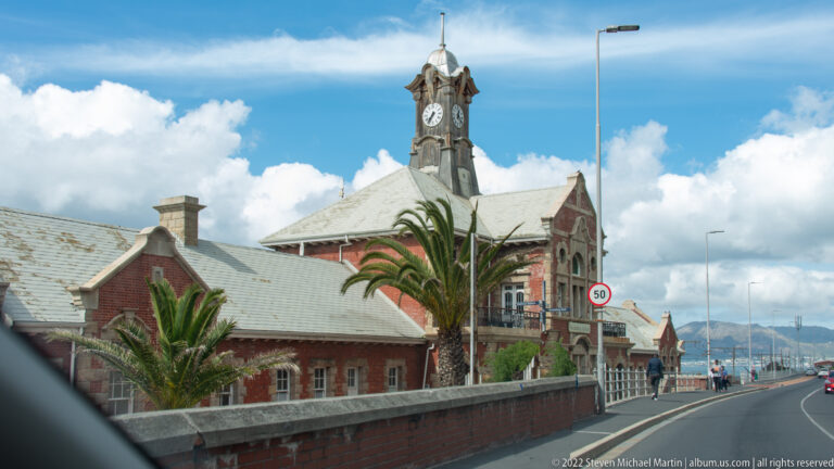 Oblique view of Muizenberg Railway Station with red-brick walls, pale roof, palm trees, and clock tower.