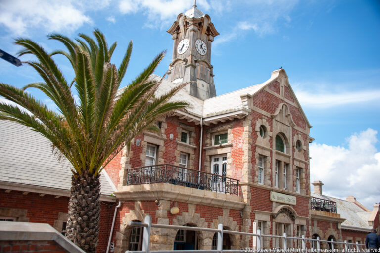 Front section of Muizenberg Railway Station with red brick, arched windows, balcony, palm tree, and clock tower.