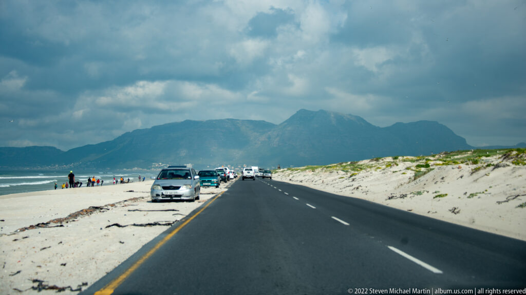 Coastal road beside a sandy beach, with parked cars, beachgoers, dunes, and mountains beneath heavy clouds.