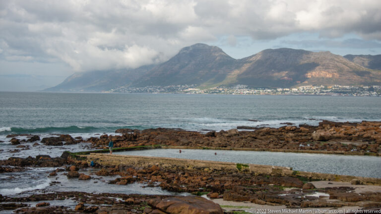 Rocky shoreline and coastal path beside False Bay, with hillside homes and cloud-covered mountains across the water.
