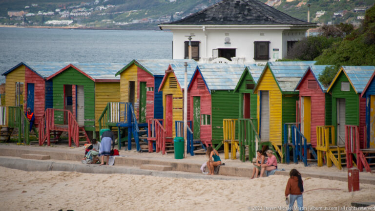Row of colorful wooden bathing boxes on Muizenberg beach with people, a white building, and bay behind.
