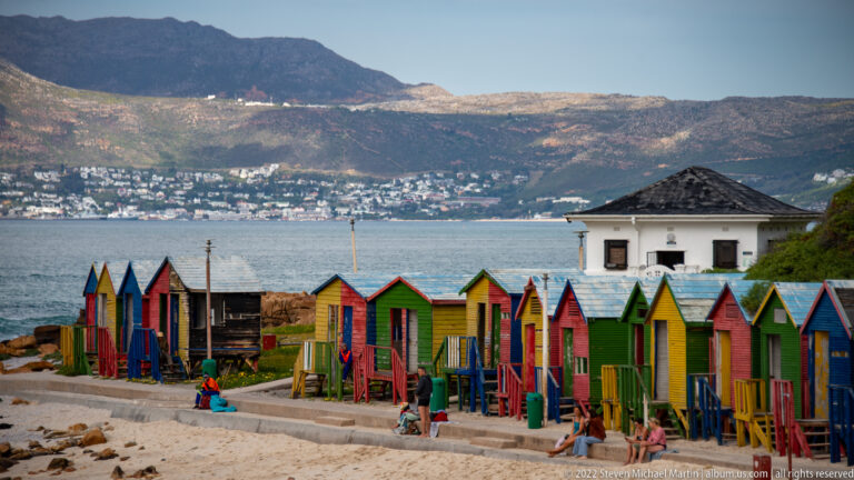 Colorful Muizenberg bathing boxes along the beach, with bay water, a white building, hillside homes, and mountains beyond.
