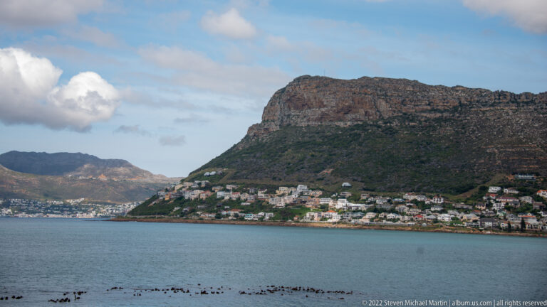 False Bay water below a steep flat-topped mountain with dense houses along its lower slopes and shoreline.