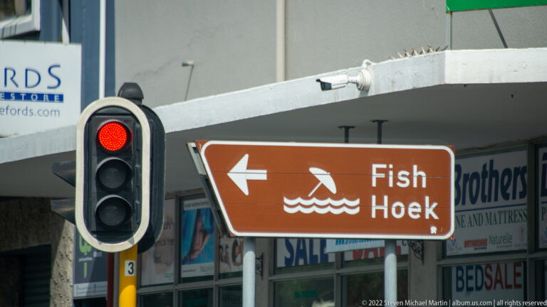 Brown Fish Hoek directional sign with swimmer symbol and left arrow beside a red traffic light.
