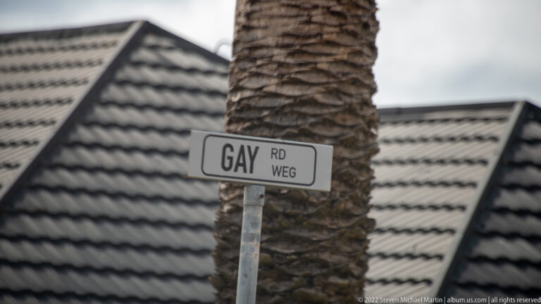 Street sign reading “GAY RD WEG” mounted in front of a palm trunk and dark tiled roof.