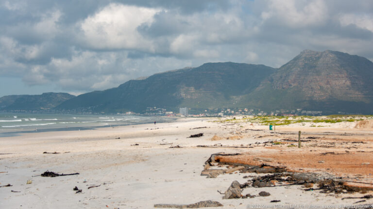 Broad sandy beach and low dunes backed by buildings and a steep mountain under broken clouds.