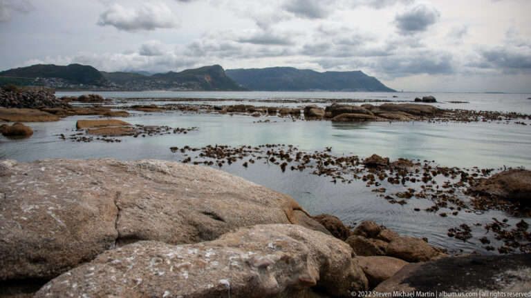 Rounded granite rocks, shallow blue water, kelp beds, and a distant mountain ridge beneath cloudy skies.