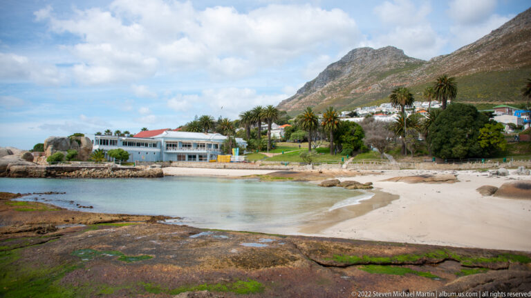 Small sandy cove with turquoise water, granite rocks, palm-lined buildings, and steep mountain slopes behind.