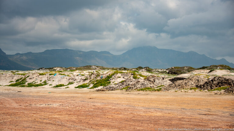 Pale vegetated dunes and reddish sandy ground in front of a dark mountain range under stormy clouds.