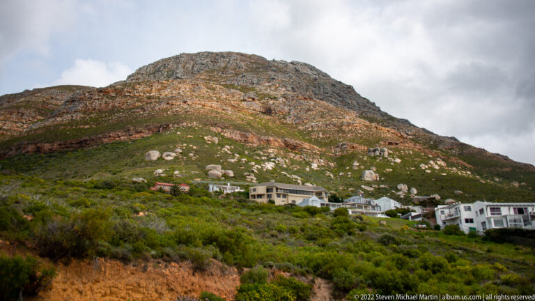 White houses beneath a rugged green mountain slope dotted with pale boulders and bands of exposed rock.