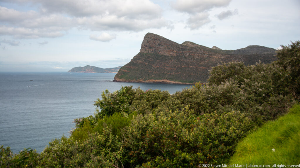 Steep rocky headland rising from blue-gray sea, with dense foreground shrubs and a smaller distant promontory.