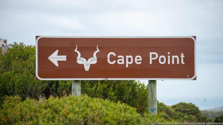 Brown directional sign with a left arrow and antelope emblem reading “Cape Point,” surrounded by coastal shrubs.