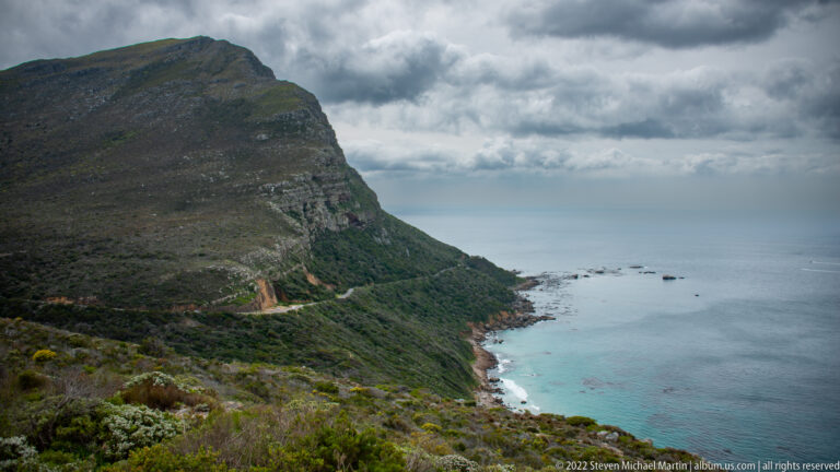 Steep dark mountain descending to a rocky shore and turquoise shallows beneath heavy clouds, with fynbos in foreground.