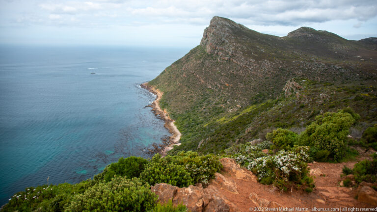 Fynbos-covered mountain ridge dropping steeply to a narrow rocky coast beside deep blue sea.