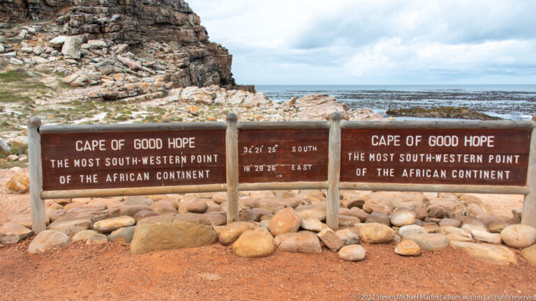 Timber Cape of Good Hope signs with coordinates on a rocky shoreline beside kelp-fringed sea.