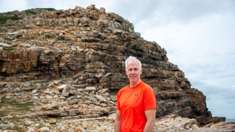 Visitor in an orange shirt standing in front of steep, fractured brown rock layers near the coast.