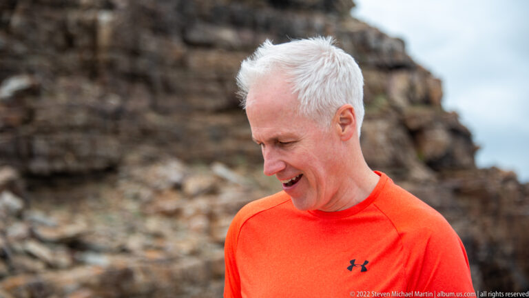 Close portrait of a visitor in an orange shirt looking downward, with a blurred rocky cliff behind.
