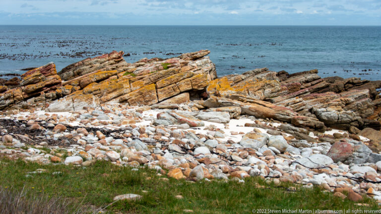 Tilted reddish and gray rock layers above a pale cobble beach, with kelp-covered sea beyond.