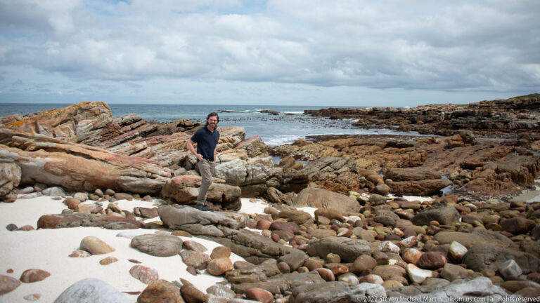 Visitor standing among rounded stones and tilted rock slabs beside tide pools and the sea under cloudy skies.