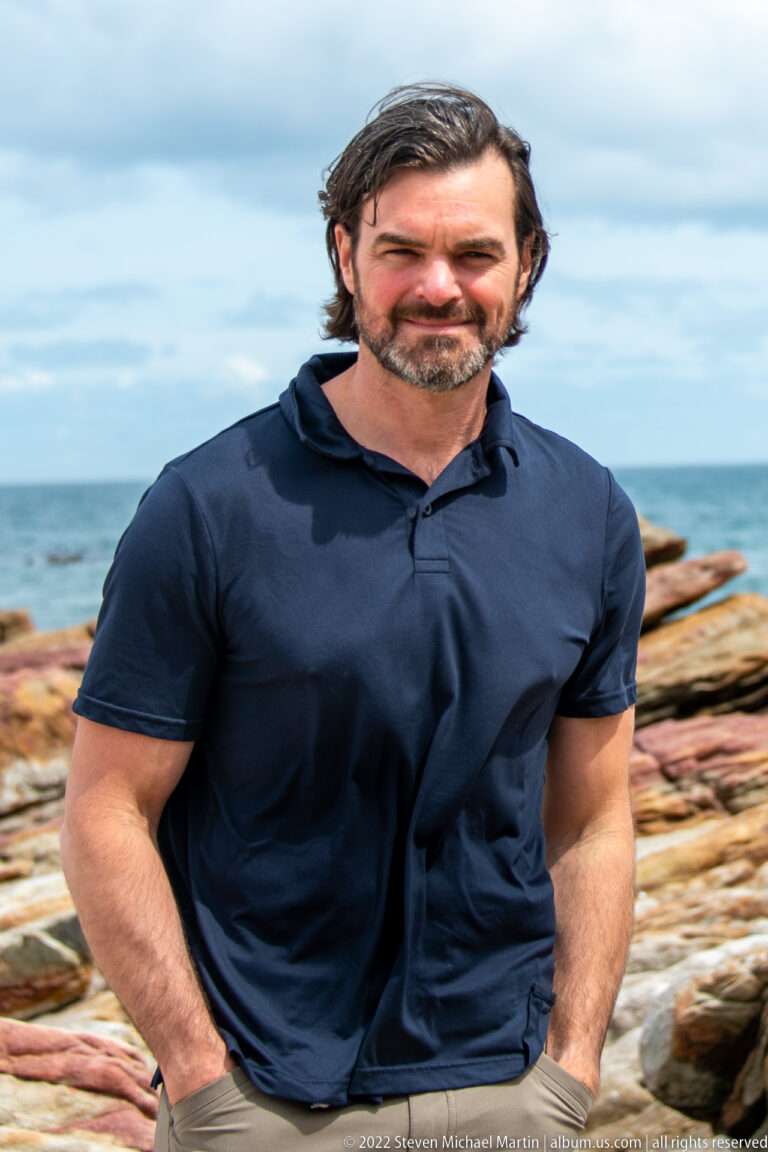 Close portrait of a visitor in a navy shirt with layered coastal rocks and sea blurred behind.
