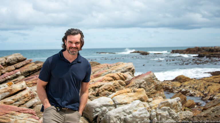 Visitor in a navy shirt standing before tilted coastal rocks, tide pools, kelp, and breaking ocean waves.