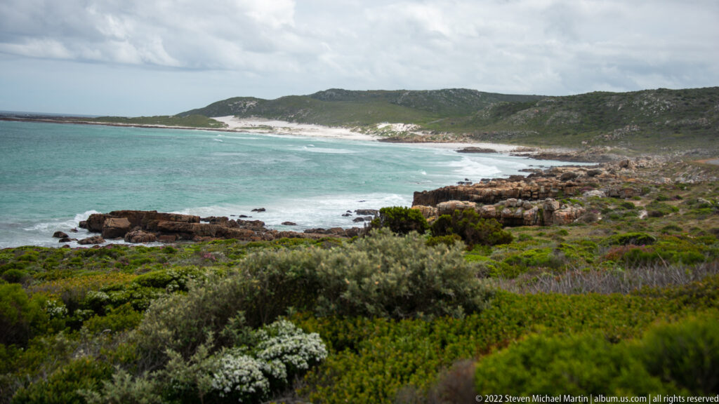 Turquoise bay curving toward a pale sandy beach, with rocky shore, dense fynbos, and low hills.