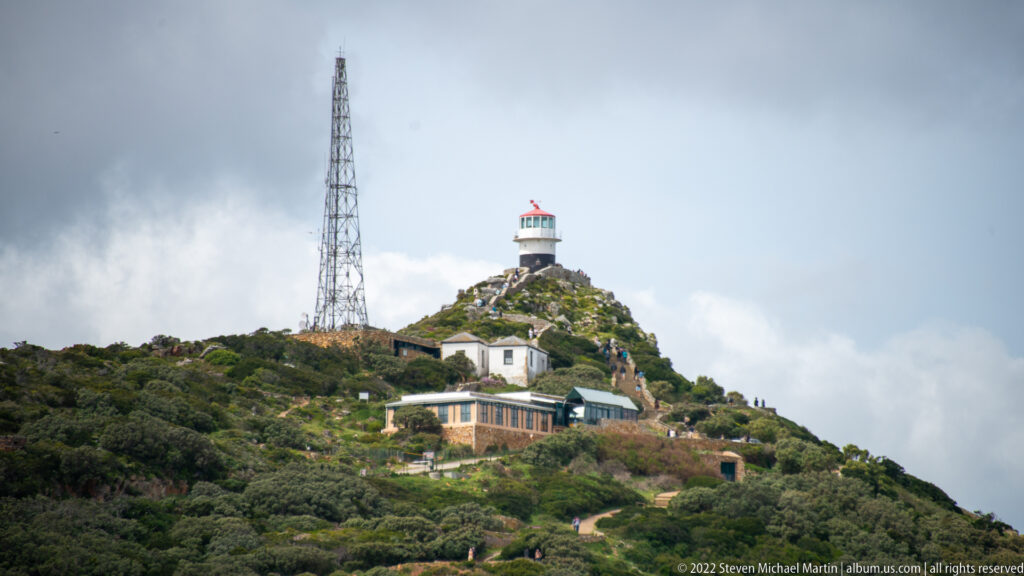 Old Cape Point Lighthouse on a shrub-covered summit beside a tall communications mast and several service buildings.