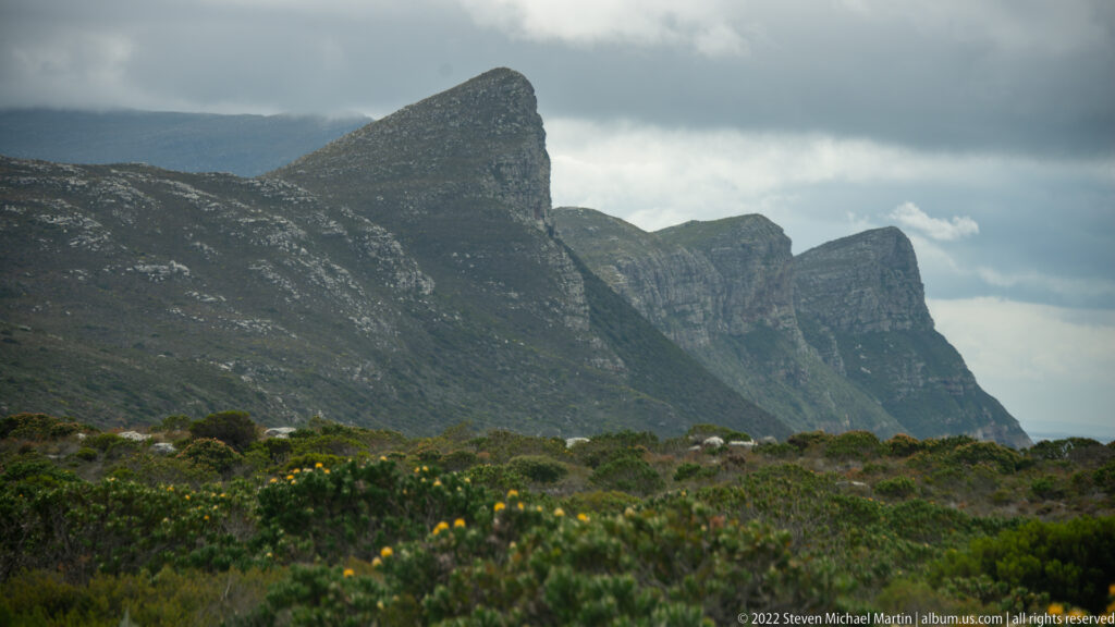Steep overlapping mountain ridges rising above dense fynbos and yellow flowers beneath heavy clouds.