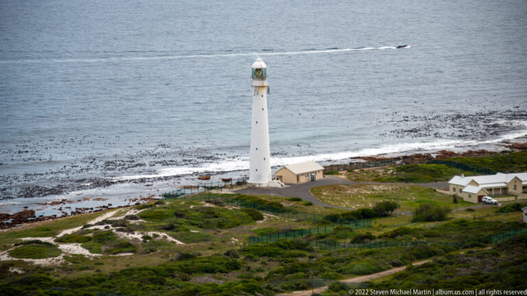 Slender white Slangkop Lighthouse beside the rocky Kommetjie shore, with service buildings, kelp beds, and Atlantic water.