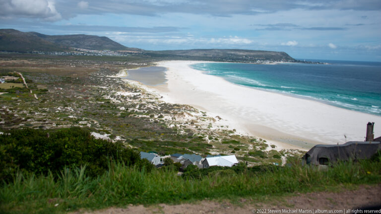 Long curving white beach beside turquoise Atlantic water, backed by dunes, low vegetation, distant houses, and mountains.