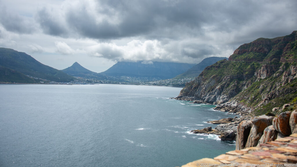 Hout Bay seen between steep mountain walls and rocky headlands under dark clouds, with settlement along the distant shore.