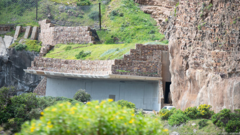 Stone retaining walls and a concrete road structure built into a steep rocky cliff with green vegetation above.