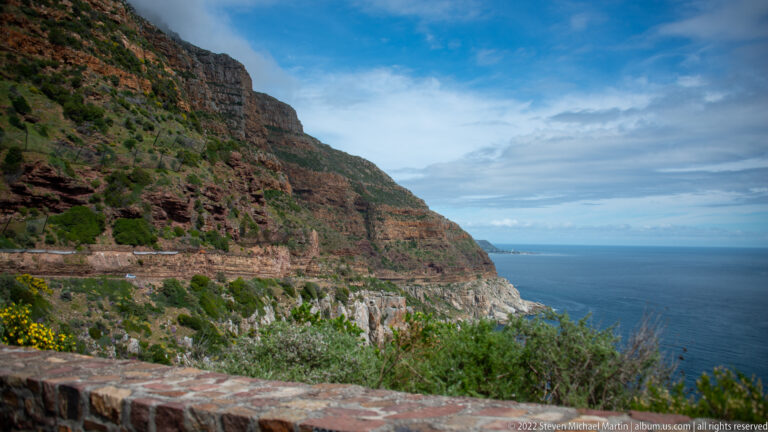 Chapman’s Peak Drive cut across a steep layered cliff above the Atlantic, with shrubs and yellow flowers in foreground.