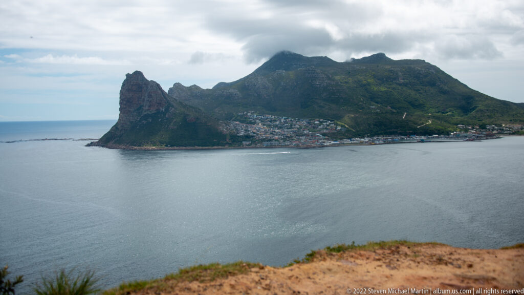 The Sentinel rising above densely settled Hout Bay, with calm gray water in foreground and cloud-covered mountains behind.