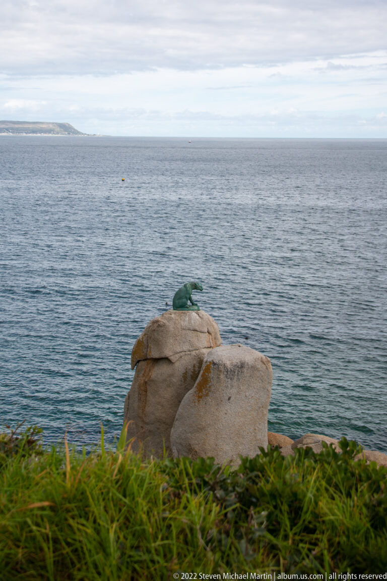 Rounded pale coastal boulder with a small green patch on top, overlooking gray-blue ocean and low vegetation.