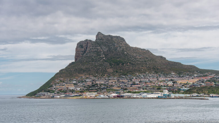 The Sentinel towering above dense Hout Bay houses and waterfront buildings, with calm bay water in the foreground.