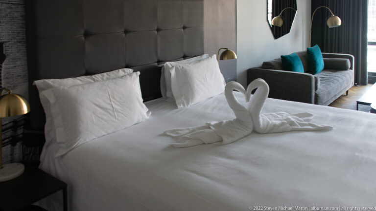 White hotel bed with two folded towel swans, tufted headboard, bedside lamps, and a small sofa near the window.