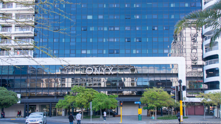 Blue-glass ONYX building with its name above the entrance, pedestrians on the sidewalk, and a palm at right.