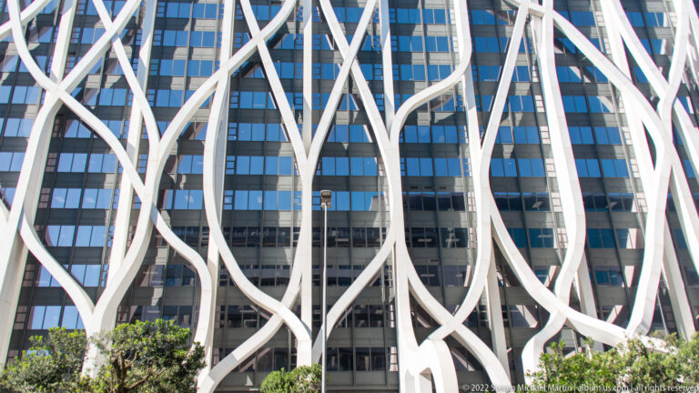 Close view of large branching white façade members crossing in front of dark blue glass windows.