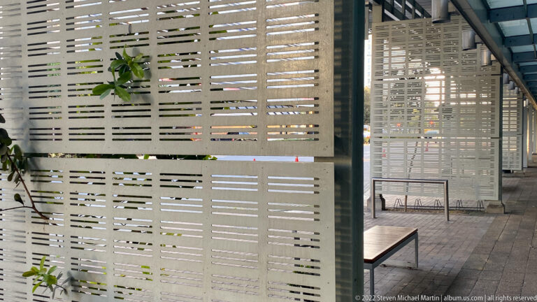 Perforated metal screen beside a paved passage, with benches, plants, glass walls, and patterned sunlight.