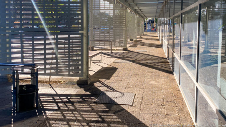 Long covered walkway with slatted screens casting geometric shadows, glass panels at right, and pedestrians in the distance.