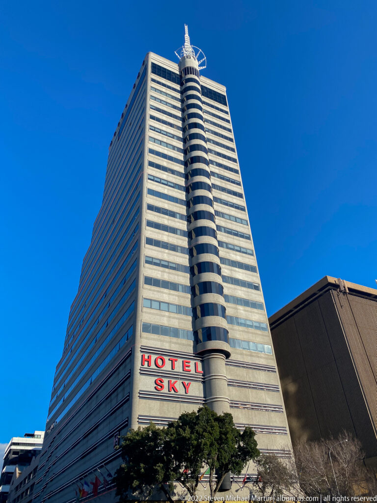 Low-angle view of the tall Hotel Sky tower with horizontal windows, rounded corner element, and red signage.